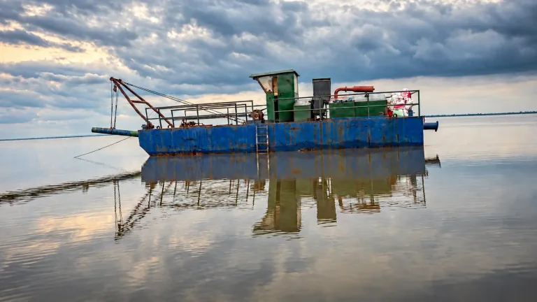 AI-edited company-supplied photo of a blue dredging vessel with a green cabin, lifting boom and reflection on calm water; deck person removed